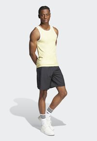 Helles gelbes Tanktop mit weißem Besatz, kombiniert mit schwarzen Shorts. Das Model trägt weiße Sneakers und schwarz-weiße gestreifte Socken. Schlichtes Design.