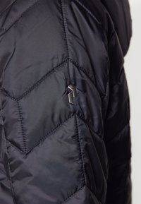 Svart boblejakke med chevron-quiltet mønster, laget av skinnende nylon, med en liten logodetalj på skulderområdet.