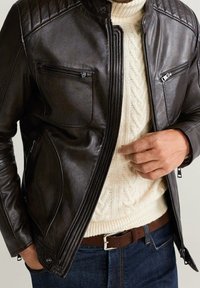 Hnědá kožená bunda s prošívanými akcenty na ramenou, dvěma zipovými kapsami na hrudi a hladkým povrchem; nošena přes krémový pletený svetr.