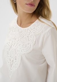Weiße Bluse mit aufwendiger Spitzenstickerei in floralen und geschwungenen Mustern, Rundhalsausschnitt und langen Ärmeln. Weicher Stofftextur.