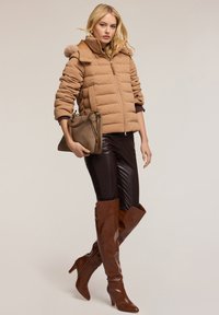Padded brown jacket z wysokim kołnierzem i futrzanym wykończeniem, w połączeniu z brązowymi skórzanymi legginsami i wysokimi butami do kolan, trzymając torebkę w tym samym stylu.