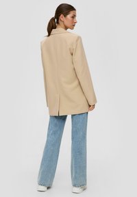 Beige taillierter Blazer mit tiefen Revers, einem einzelnen Rückenschlitz und leicht übergroßer Passform, kombiniert mit hellblauen Schlagjeans und weißen Schuhen.