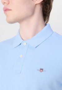 Világoskék pólóing, gallérral, két gombos záródással. Az embroidery logo a bal mellen pajzs mintát mutat.