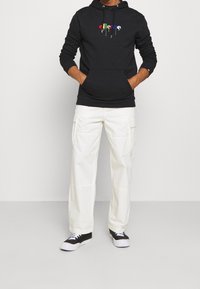 Sweat à capuche noir avec logo coloré, poche avant kangourou ; associé à un pantalon cargo blanc et des baskets noires avec accents blancs.