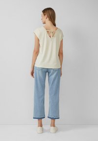 Vrouw in een crèmekleurige top met korte mouwen en een strikdetail op de rug, lichtblauwe cropped wijde jeans en witte sneakers, staande tegen een effen muur.