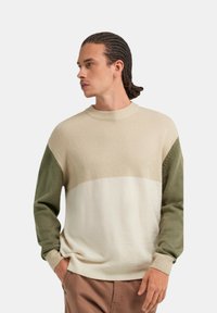 Maglione in maglia con scollo a girocollo, caratterizzato da blocchi di colore beige, bianco e verde oliva. La texture appare morbida e lavorata.