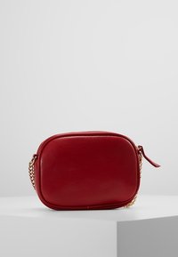 Czerwona skórzana torebka typu crossbody o zaokrąglonym kształcie, ze złotym łańcuszkiem i zamkiem błyskawicznym. Gładka tekstura z przeszyciami na brzegach.