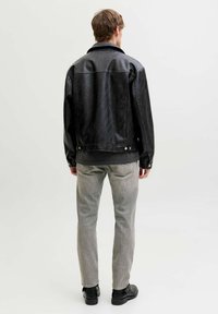 Chaqueta de cuero negra con textura lisa, que presenta un cuello puntiagudo y botones a presión. Llevada sobre jeans gris claro y botas negras.