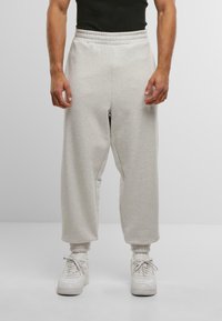 Muž oblečený v svetlošedých širok�ých teplákoch, bielych ponožkách a bielych teniskách na neutrálnom pozadí, s uvoľnenými rukami po stranách.