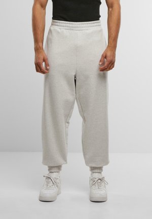 Mann trägt hellgraue, weit geschnittene Sweatpants, weiße Socken und weiße Sneaker vor einem neutralen Hintergrund, die Hände entspannt an den Seiten.