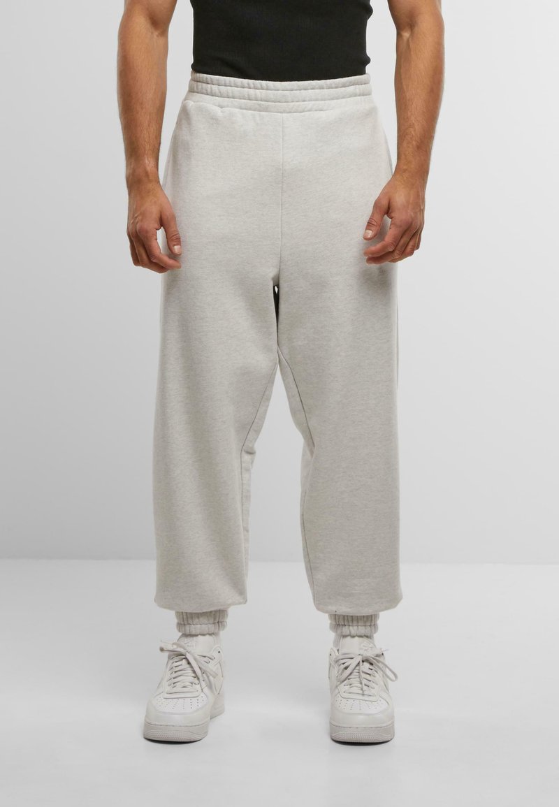 Muž oblečený v svetlošedých širokých teplákoch, bielych ponožkách a bielych teniskách na neutrálnom pozadí, s uvoľnenými rukami po stranách.