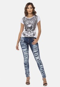 Witte graphic t-shirt met zwart schedel- en vleugeldesign, gecombineerd met versleten blauwe skinny jeans met grote scheuren en rafelige randen.