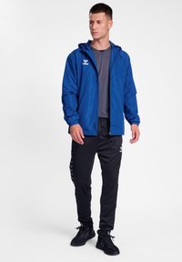 Blå lättviktsjacka med huva, som bärs tillsammans med en grå skjorta och svarta träningbyxor med sidostycken. Kombineras med svarta sneakers.
