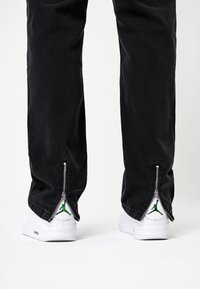 Czarne dżinsy z zamkami błyskawicznymi na kostkach, w połączeniu z białymi sneakersami z zielonym logo Jumpman, podkreślającymi kontrast i detale.