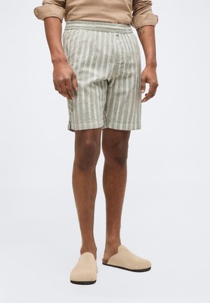 Άνδρας που φοράει μπεζ παπούτσια χωρίς κορδόνια, ανοιχτοπράσινα και λευκά κάθετα ριγέ σορτς και μπεζ πουκάμισο με μακριά μανίκια που είναι σηκωμένα.