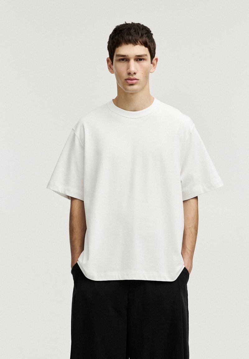 Tricou alb din bumbac, cu o croială lejeră, mâneci scurte și guler rotund. Textură netedă, design minimalist. Asortat cu pantaloni scurți negri.