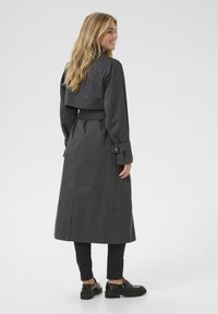 Lungo cappotto nero con vita cintata, polsini abbottonati e schiena ventilata. Indossato sopra pantaloni neri e scarpe nere chunky.