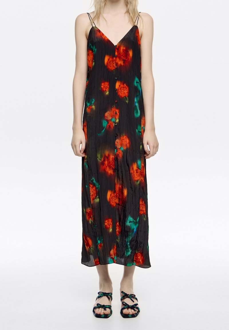 Robe longue noire sans manches à boutons, avec un motif floral rouge et vert, portée avec des sandales noires à brides.