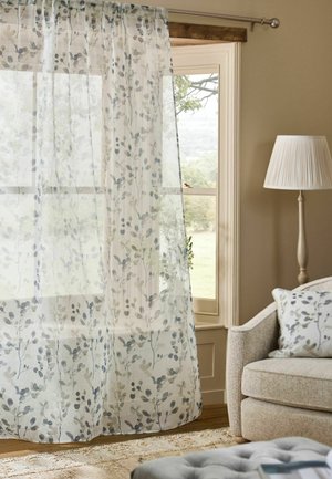 Transparente weiße Vorhänge mit grauen Blattmustern hängen an einem Fenster neben einem beigen Sessel mit passendem blattgemustertem Kissen und einer Stehlampe.