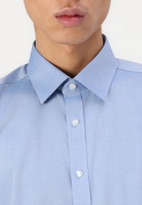 Cămășă albastru deschis cu guler, realizată dintr-un material neted, are un sistem de închidere cu nasturi albi și un design clasic al gulerului.