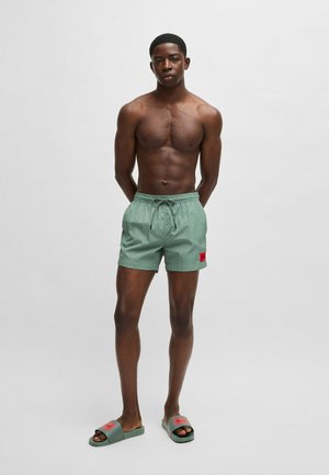 Pantaloncini da bagno verdi con vita elastica e cordino, con una toppa rossa del logo sul lato sinistro; indossati con ciabatte verdi coordinate.