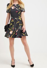 Robe noire à imprimé floral avec une jupe évasée, des manches courtes et un corsage ajusté. Portée avec des talons noirs et une pochette.