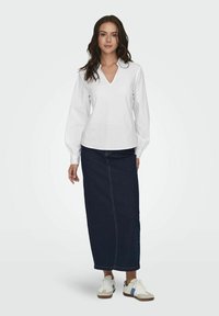 Blusa bianca a maniche lunghe con scollo a V e maniche a sbuffo, abbinata a una maxi gonna di jeans scura e sneakers bianche con dettagli blu.