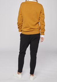 Mörkgul långärmad tröja med rundad halsringning och ribbade ärmslut, kombinerad med svarta smalrandiga byxor och vita sneakers. Slät tygkänsla.