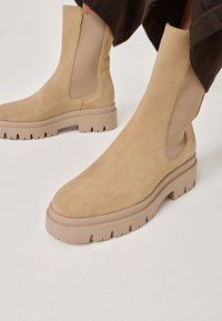 Beige semišové kotníkové boty s elastickými bočními panely a robustní gumovou podrážkou s výrazným vzorem.