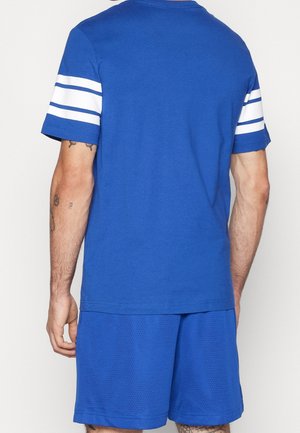Mann trägt blaues kurzärmeliges Hemd mit zwei weißen Streifen auf jedem Ärmel und blaue Netzshorts, der von der Kamera weg schaut.