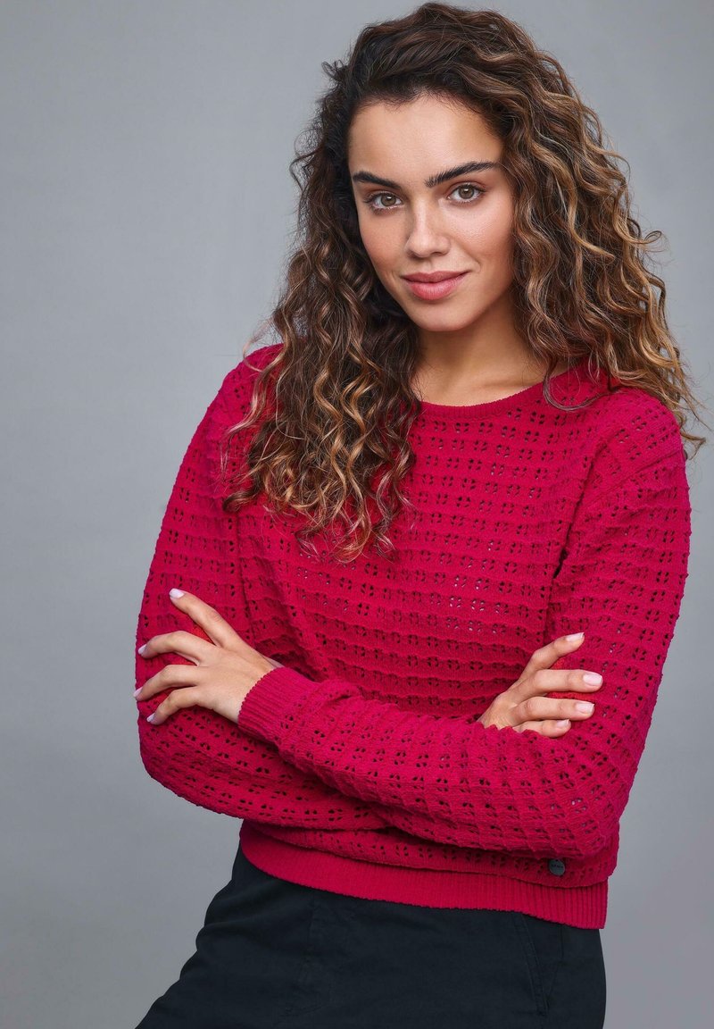 Czerwony sweter z dzianiny, z teksturowanym wzorem, ściągaczami przy mankietach i u dołu. Model pozuje z założonymi rękami na tle w neutralnych kolorach.