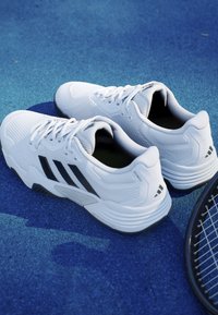 Pár bielych športových topánok Adidas s čiernymi pruhmi na modrej povrchovej ploche kurtu vedľa tenisovej rakety.
