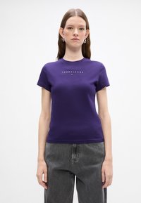 Violeta kokvilnas T-krekls ar apaļu kakla izgriezumu un īsām piedurknēm, ar baltu "TOMMY JEANS" logo priekšā un pieguļošu dizainu.