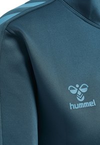 Abbigliamento sportivo Hummel in tessuto verde scuro, caratterizzato da una texture liscia, accenti azzurri sulle spalle e un logo prominente sul petto.