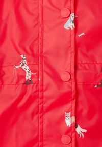 Röd jacka med hundillustrationer. Slätt, glänsande material med knäppning och stickade sömmar. Hundar i olika positurer och färger.