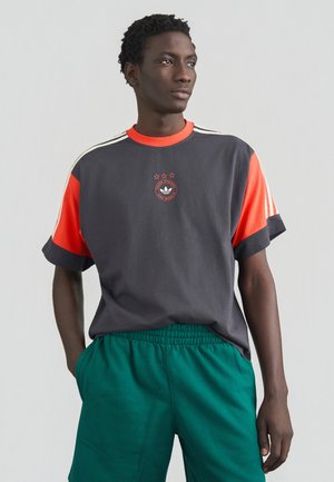 Hall ja oranž T-särk ümmarguse kaelusega, kolme tähe graafika ja Adidas logo; sobitatud roheliste pükstega, millel on elastne vöökoht.