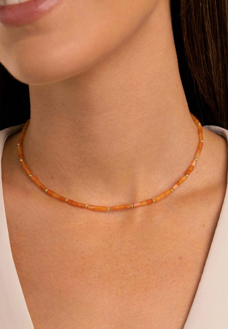 Collier avec de petites perles rectangulaires orange séparées par de minuscules perles dorées, porté autour du cou d'une femme avec un haut clair.