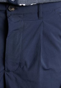 Pantalones azul marino con frente plisado, textura suave, costuras visibles y cierre de botón en la cintura.