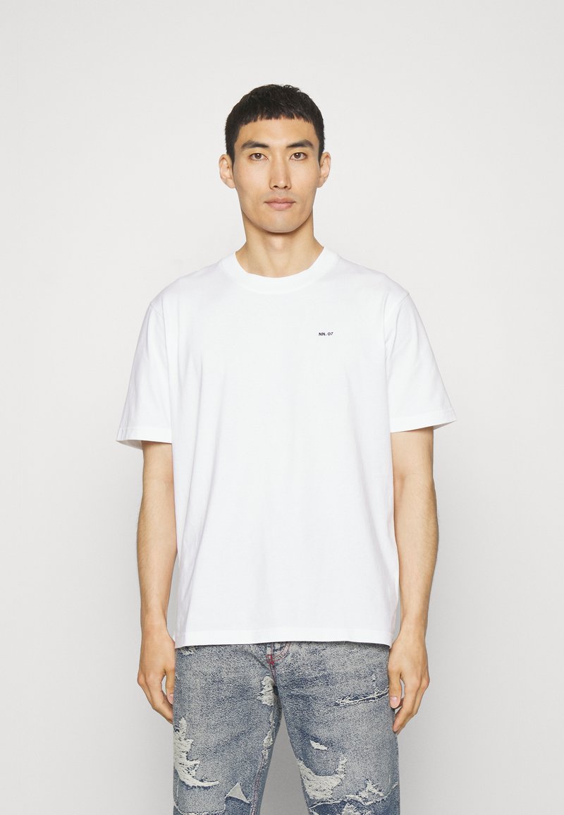 Weißes Baumwoll-T-Shirt mit Rundhalsausschnitt und kurzen Ärmeln, mit einem kleinen Logo auf der linken Brust. Kombiniert mit destroyierten Jeans.
