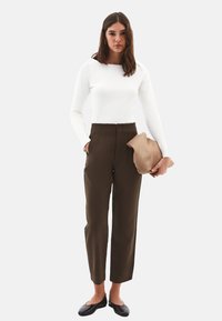 Kvinde i hvid langærmet top og højtaljede brune bukser holder en tan clutch i den ene hånd, mens hun har den anden hånd i lommen, iført sorte flade sko.