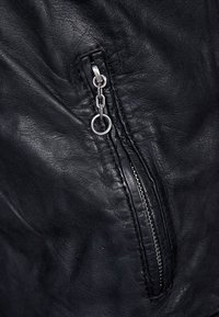 Gros plan sur une veste en cuir noir avec une poche zippée dotée d'un onglet en argent et de détails en chaîne. Le cuir a une finition texturée.