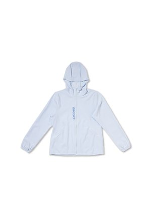 Mujer con chaqueta azul claro con cremallera y logo "BROOKS", pantalones oscuros, sonriendo y mirando hacia su izquierda sobre un fondo liso.