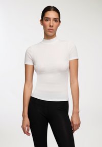 Biała prążkowana bluzka z krótkim rękawem i wysokim dekoltem, dopasowany krój podkreślający sylwetkę, w połączeniu z czarnymi legginsami. Gładka faktura całości.