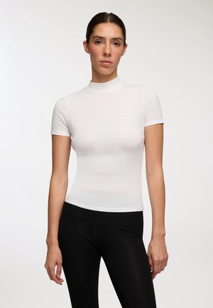 Top bianco a maniche corte, con collo alto e design aderente che accentua la forma, abbinato a leggings neri. Texture liscia in generale.
