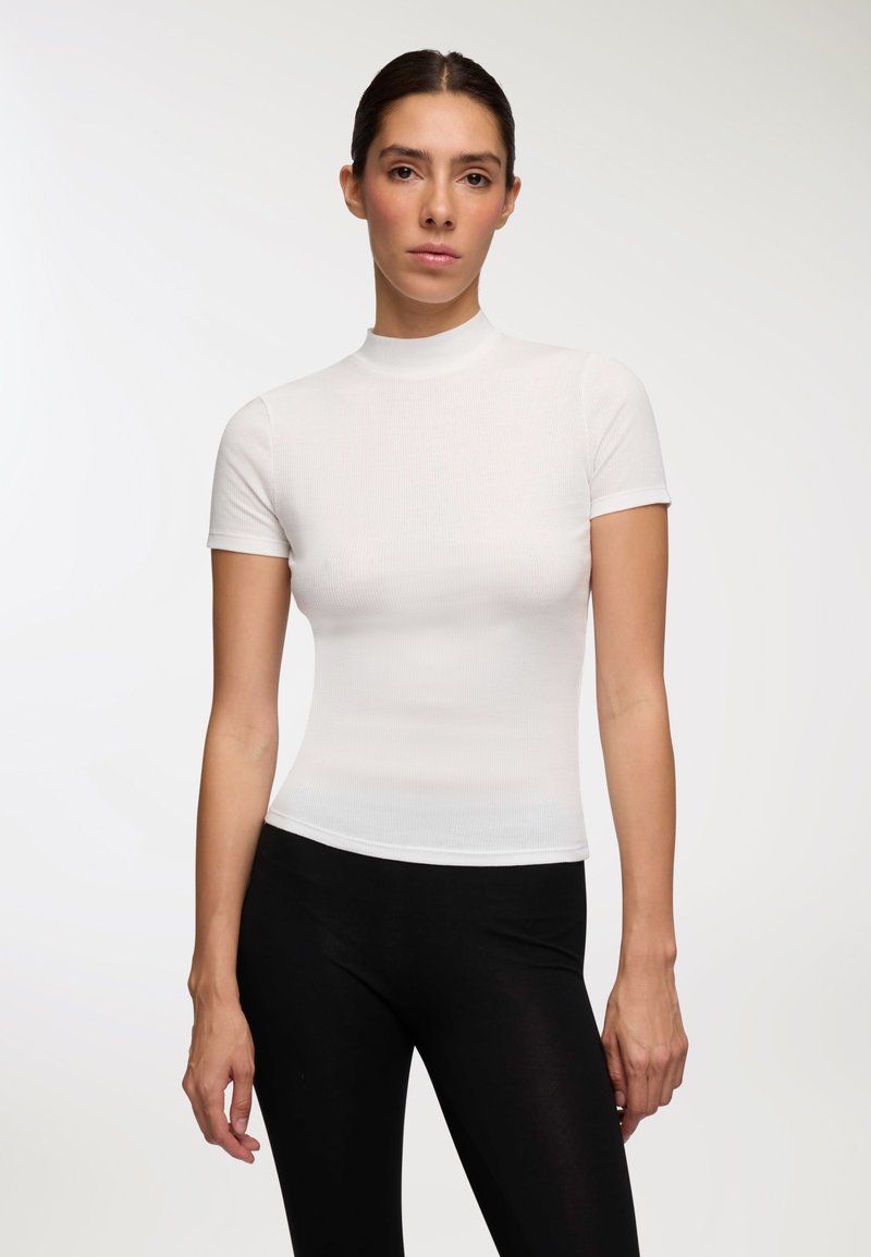 Biała prążkowana bluzka z krótkim rękawem i wysokim dekoltem, dopasowany krój podkreślający sylwetkę, w połączeniu z czarnymi legginsami. Gładka faktura całości.