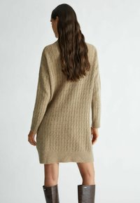 Abito maglione in beige chiaro con texture a costine e maniche lunghe. Arriva sopra il ginocchio, abbinato a stivali alti lucidi marroni.