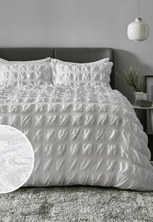 King-Size-Bett mit strukturiertem weißem Steppbett und passenden Kissen, dunkelgrauem Kopfteil, grauer Wand, hängender weißer Lampe und kleinem Pflänzchen auf dem Beistelltisch.