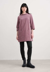 Donna che indossa una tunica a maniche lunghe mauve, pantaloni neri aderenti e anfibi neri, in piedi contro uno sfondo bianco semplice.