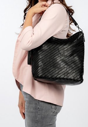 Kvinde iført lyserød blazer og grå jeans bærer en stor sort lærredtaske med diagonalt mønster.