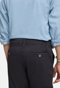 Pantalones oscuros de color marino con bolsillos traseros. Presentan botones dorados y detalles de costura. Combinados con una camisa de manga larga de color azul claro.